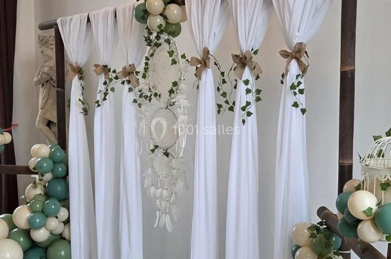 Rideau blanc décoré de feuillage, attrape-rêves et nœuds en toile de jute, entouré de ballons verts et blancs.