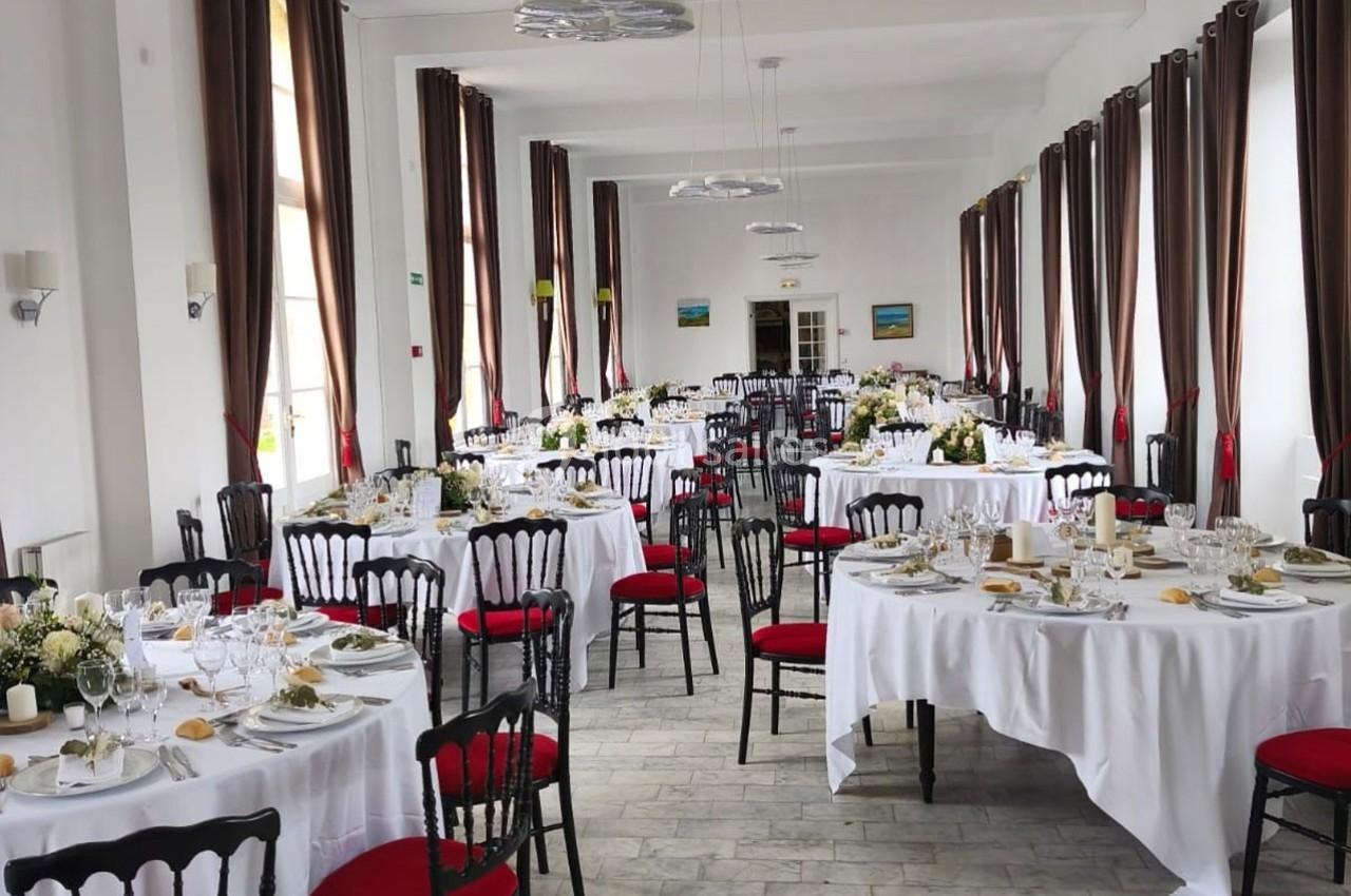 Salle de réception lumineuse avec des tables rondes dressées, nappes blanches, chaises noires à assise rouge.