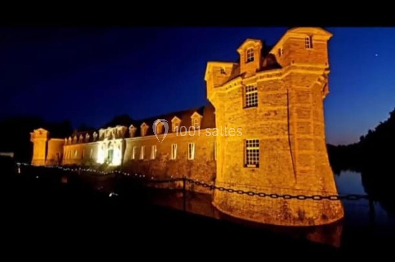 Château illuminé de nuit avec des reflets sur l'eau, entouré d'une chaîne en premier plan.