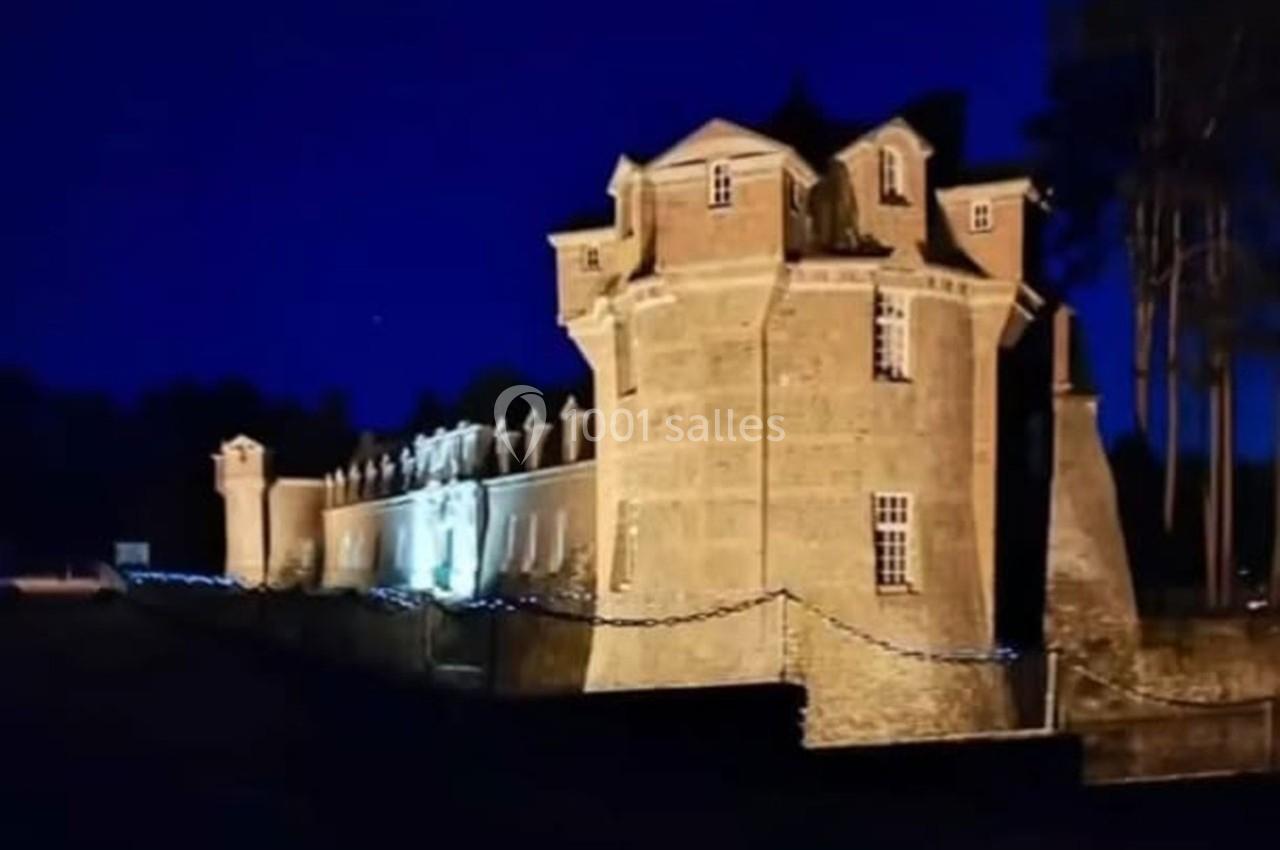 Château illuminé de nuit avec des murs en pierre, entouré d'arbres et d'une chaîne métallique au premier plan.
