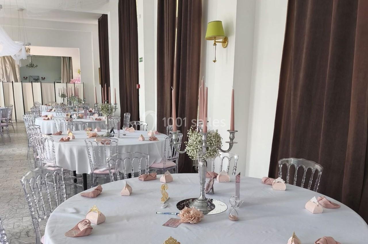 Salle de réception décorée avec des tables rondes, nappes blanches, bougies roses et pliages de serviettes élégants.