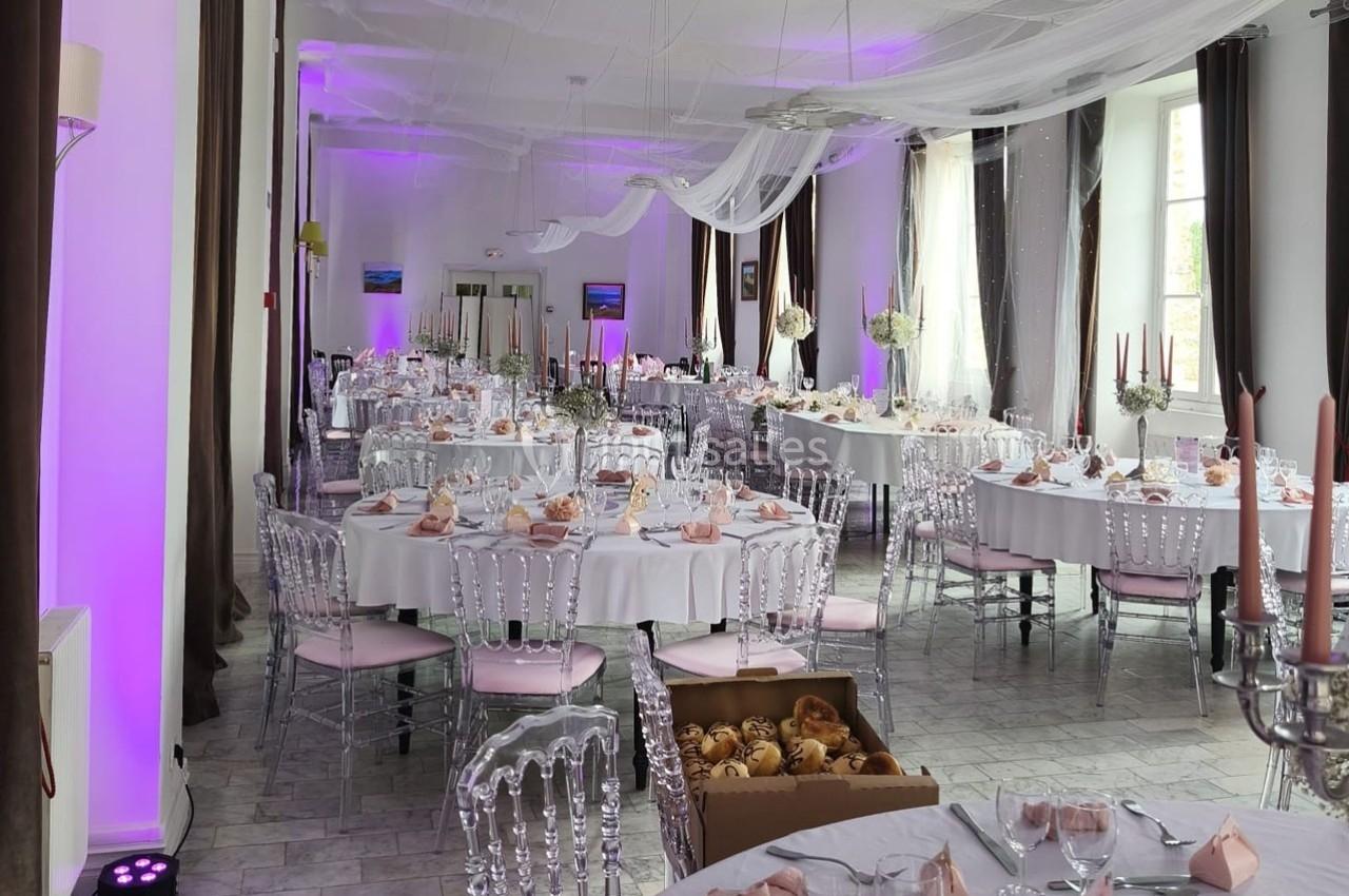 Salle de réception décorée avec des tables rondes, nappes blanches, chaises transparentes et éclairage violet.