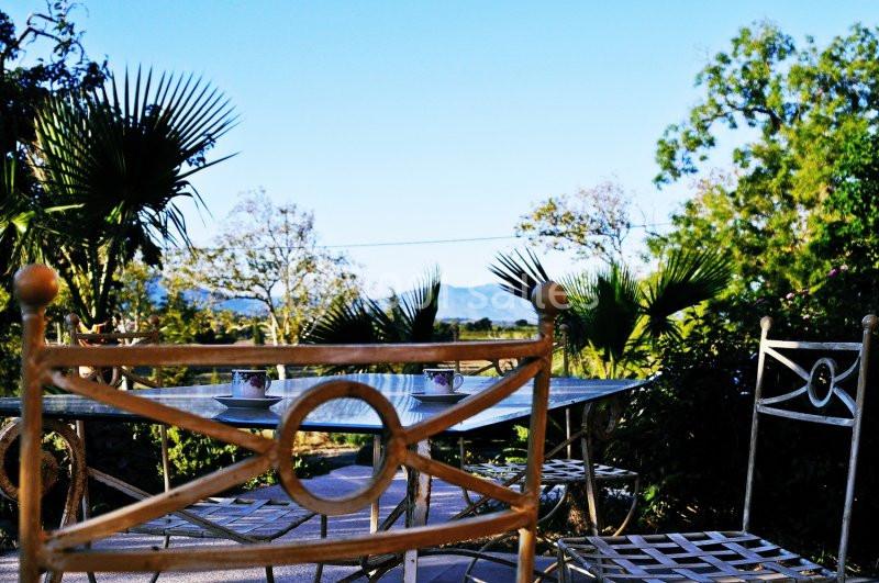 Table en fer forgé avec tasses à café sur une terrasse, entourée de végétation et vue sur des collines au loin.