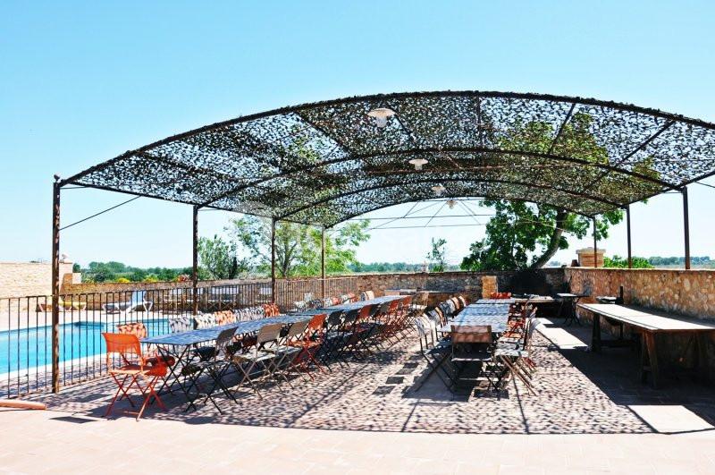 Terrasse extérieure ombragée avec de longues tables et chaises, surplombant une piscine et un paysage rural.