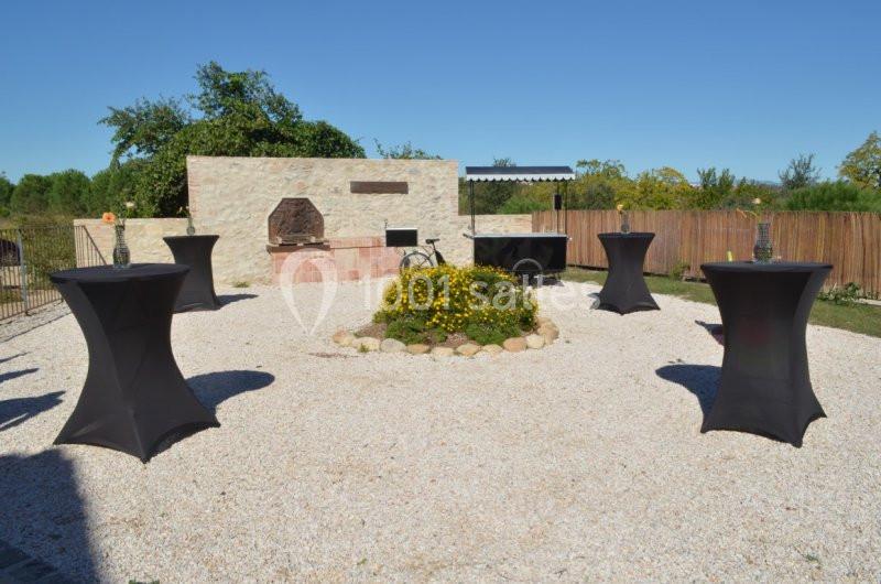 Espace extérieur avec tables hautes noires, barbecue en pierre et parterre de fleurs au centre, sous un ciel dégagé.