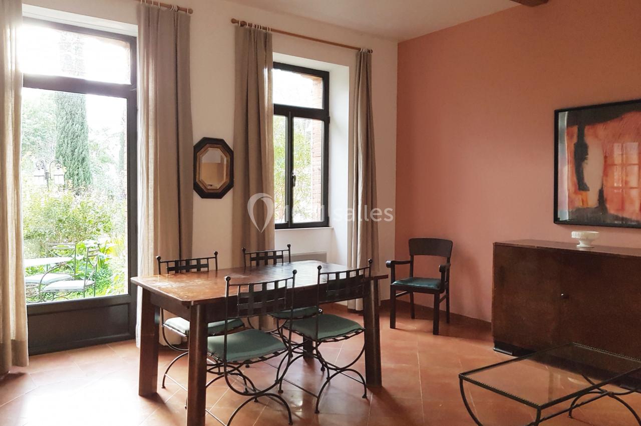 Salle à manger lumineuse avec table en bois, chaises en métal, murs roses, grandes fenêtres et vue sur un jardin.