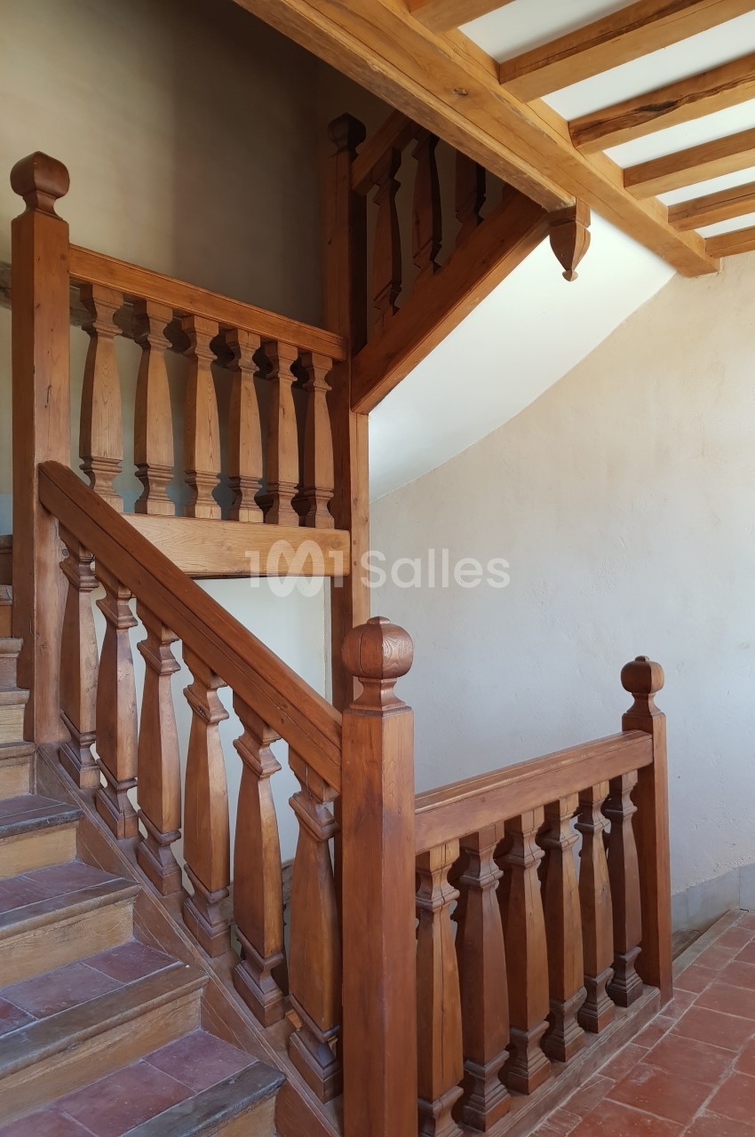 Escalier en bois avec balustrades sculptées, situé dans un intérieur aux murs clairs et poutres apparentes.