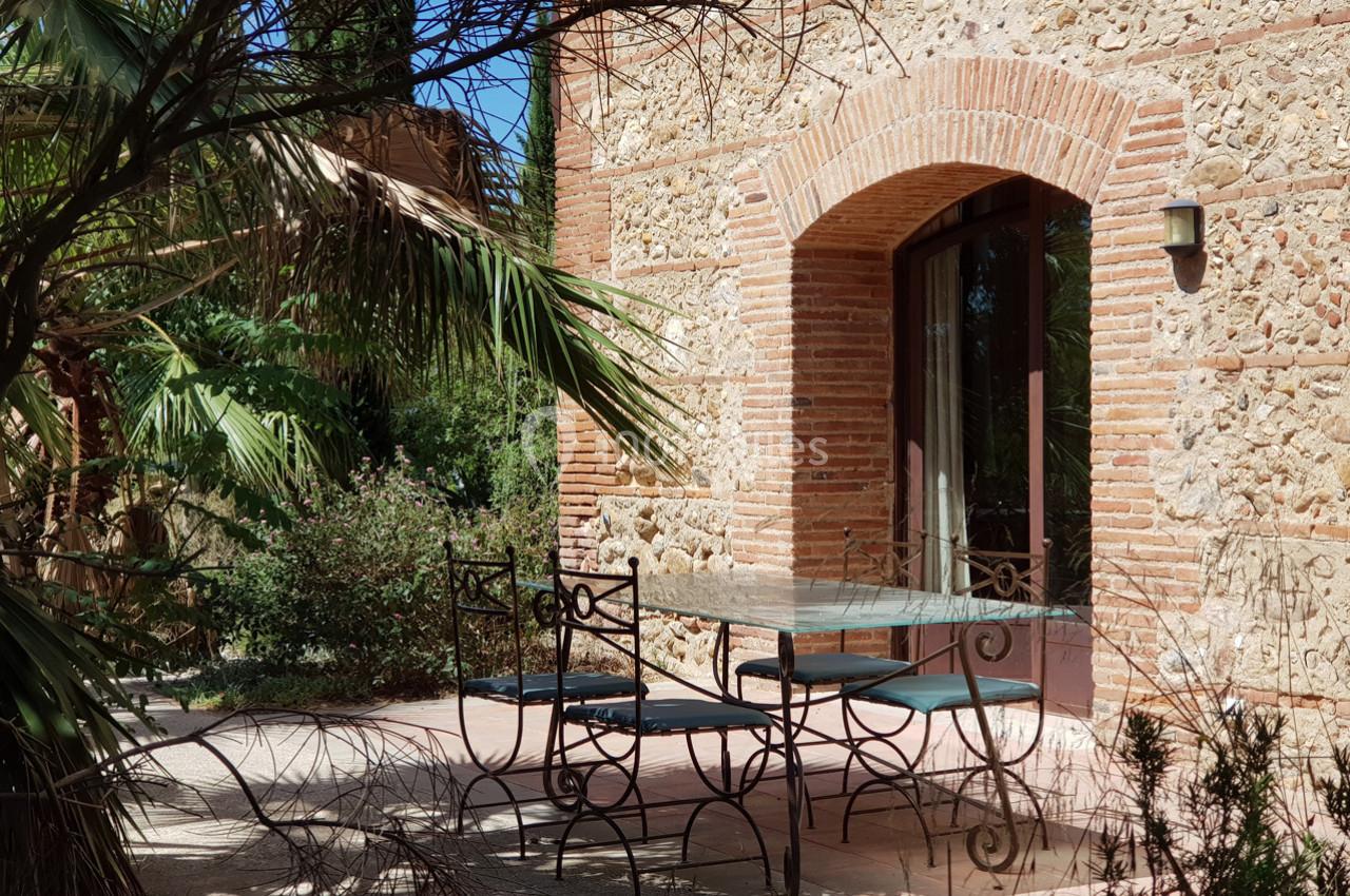 Table et chaises en fer forgé sur une terrasse ensoleillée, devant une façade en pierre avec une grande porte vitrée.