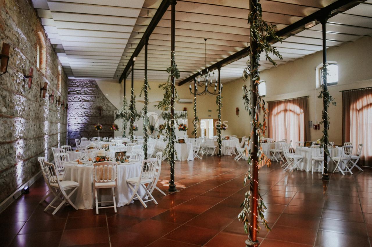 Salle de réception lumineuse avec tables rondes dressées, chaises blanches et décorations végétales suspendues.