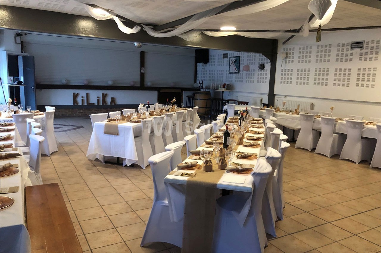 Salle de réception décorée avec des tables alignées, nappes blanches et chemins de table beige, prête pour un événement.