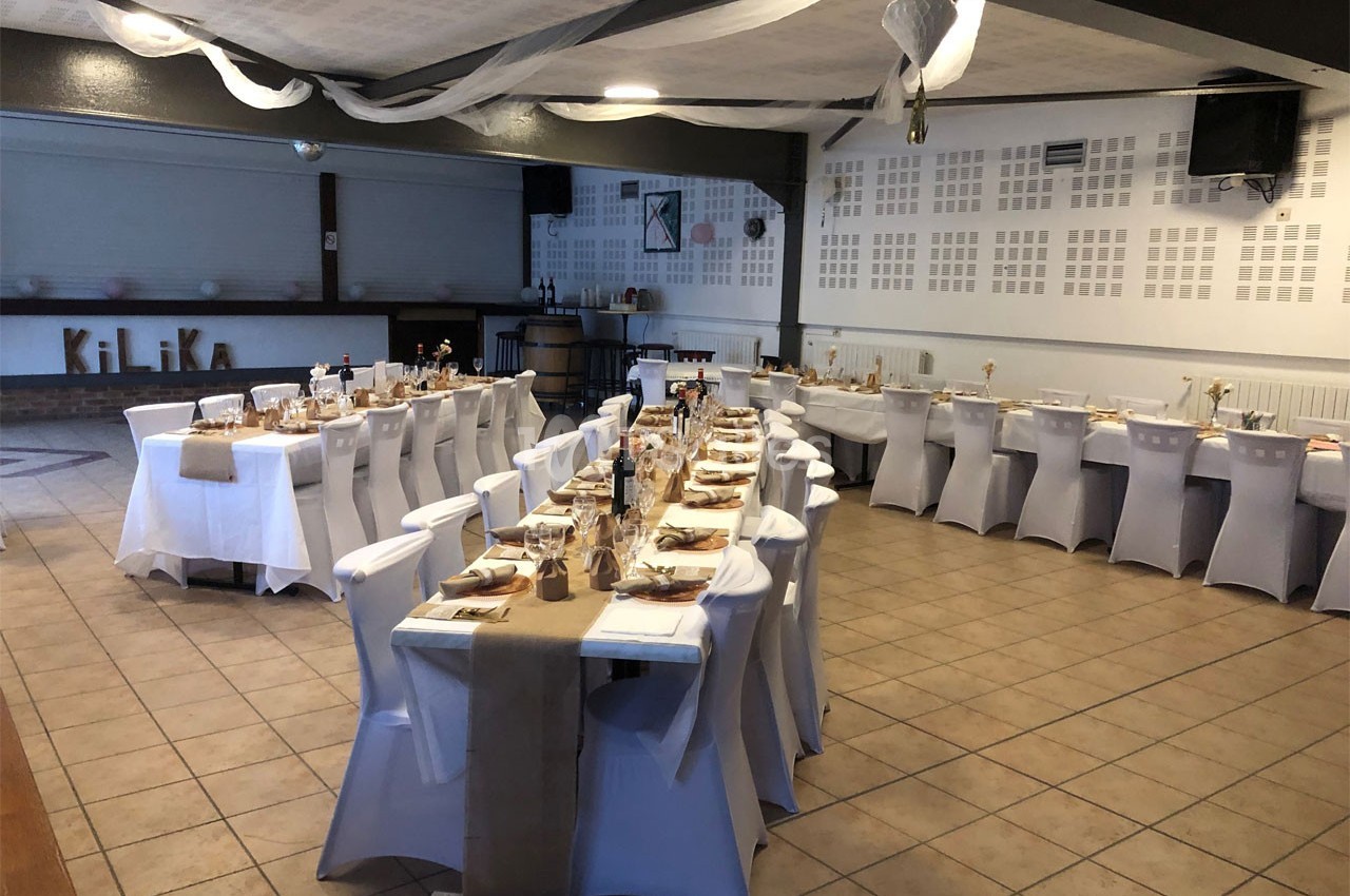 Salle de réception décorée avec des tables alignées, nappes blanches et chemins de table en tissu beige.