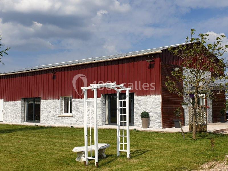 Bâtiment à façade rouge et blanche avec une pergola blanche et un banc en pierre dans un jardin verdoyant.