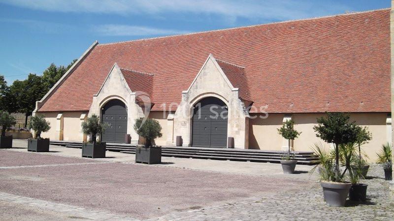 Bâtiment avec une grande toiture en tuiles rouges, deux portes voûtées et une cour pavée bordée de plantes en pots.
