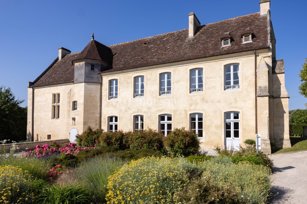 Façade d'un bâtiment ancien en pierre avec toit en ardoise, entouré d'un jardin fleuri sous un ciel bleu.