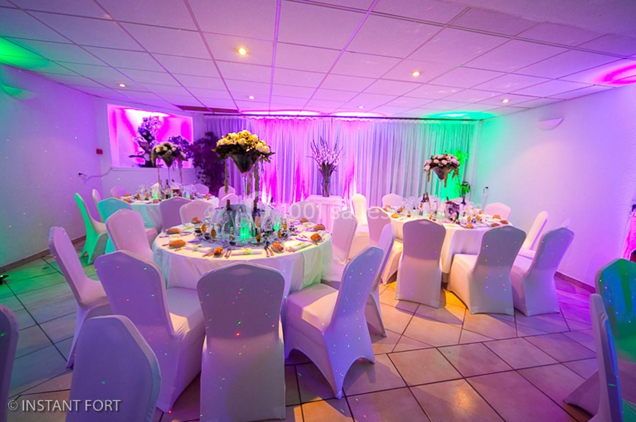 Salle de réception décorée avec des tables rondes dressées, chaises blanches et éclairage coloré.