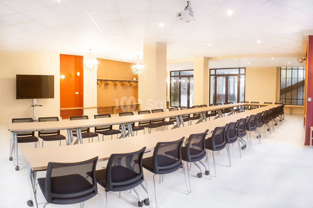 Salle de réunion lumineuse avec de longues tables, des chaises noires, un écran et des lustres modernes.