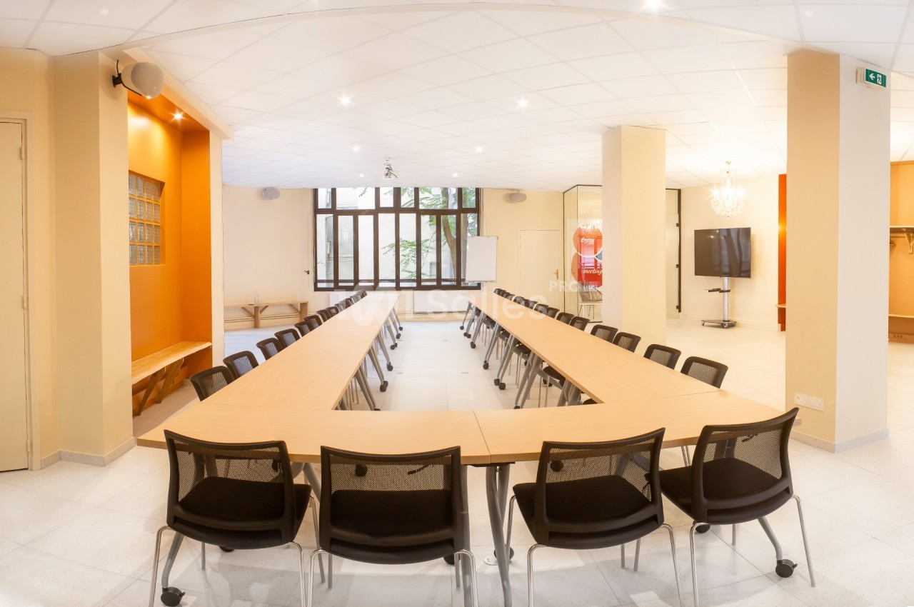 Salle de réunion lumineuse avec tables disposées en U, chaises noires, tableau blanc et écran au fond.