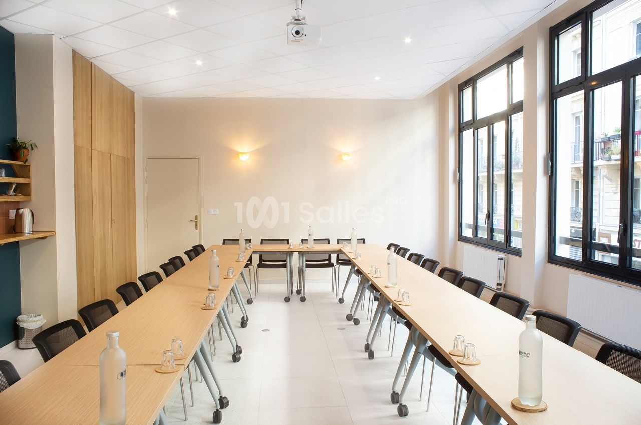 Salle de réunion lumineuse avec grandes fenêtres, tables disposées en U, chaises noires et bouteilles d'eau sur les tables.