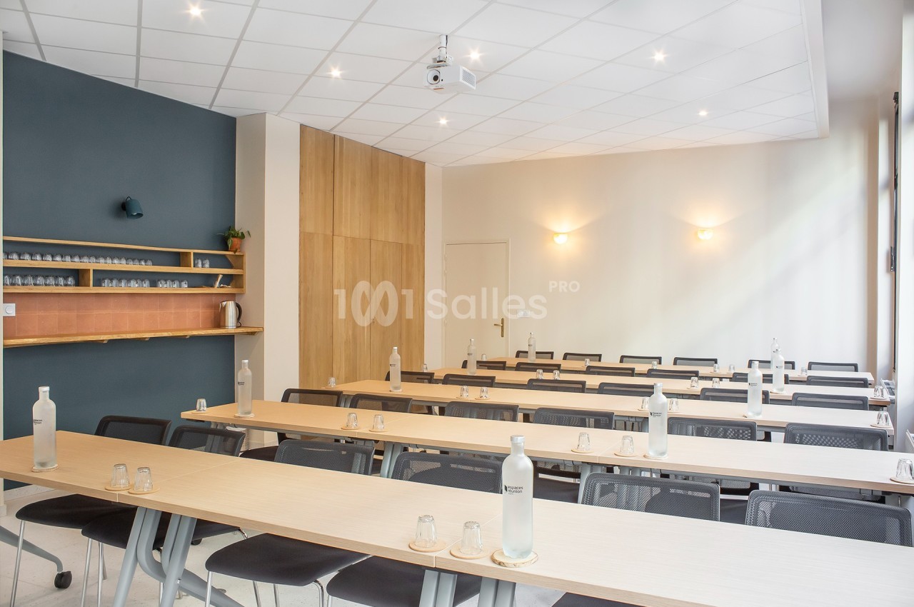 Salle de réunion lumineuse avec des tables alignées, des chaises noires et des bouteilles d'eau disposées.