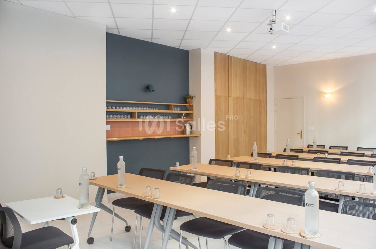 Salle de réunion lumineuse avec tables en bois, chaises noires, bouteilles d'eau et étagères murales en arrière-plan.