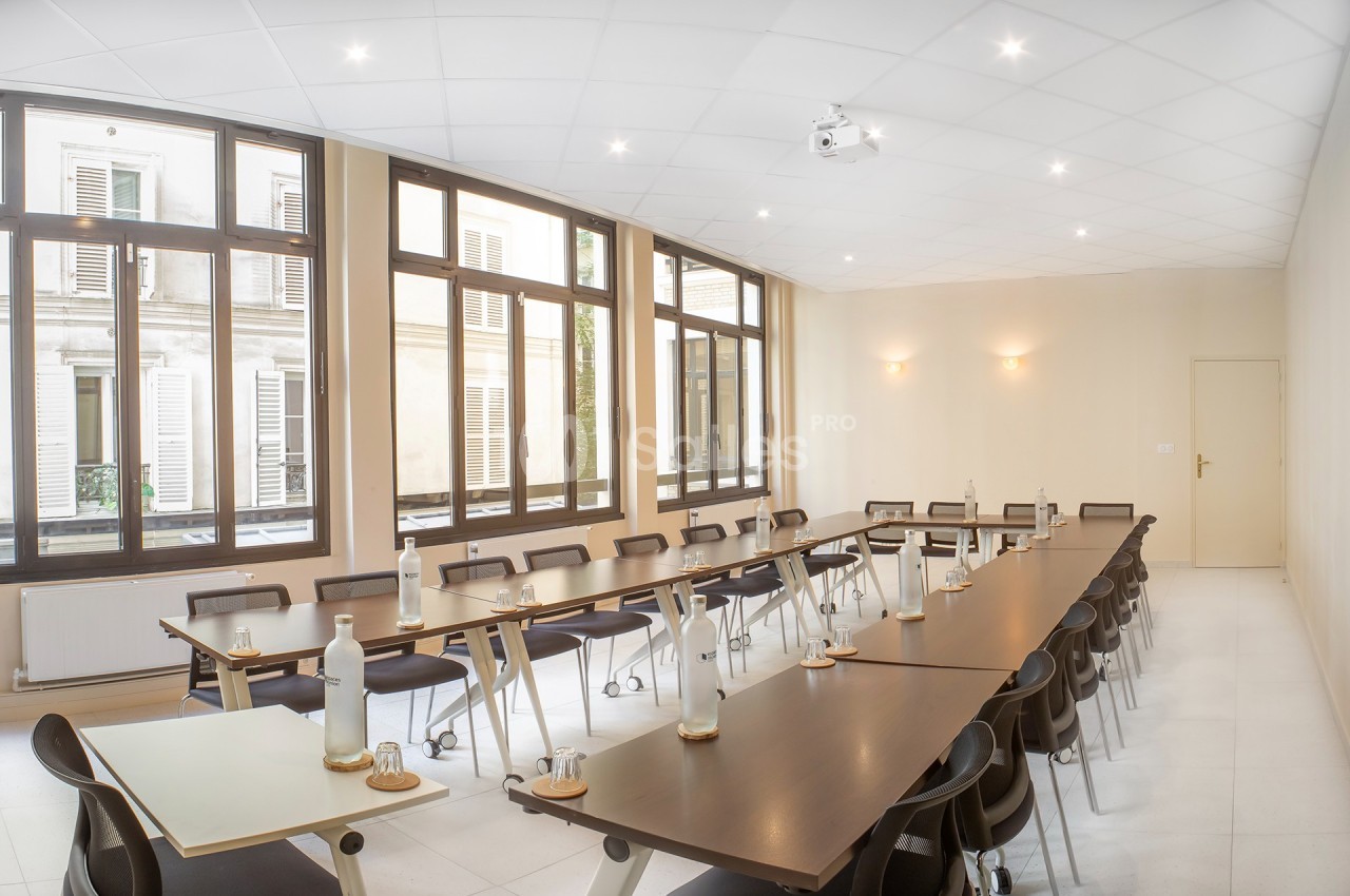 Salle de réunion lumineuse avec grandes fenêtres, tables en bois disposées en U et chaises noires.