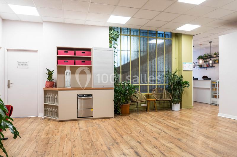 Espace intérieur lumineux avec sol en bois, plantes, chaises en rotin et étagères contenant des boîtes roses.