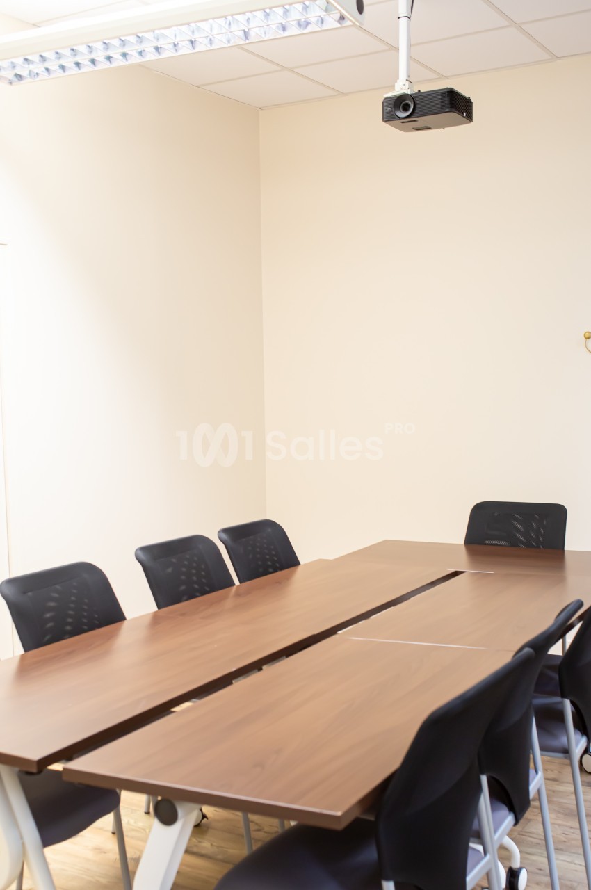 Salle de réunion lumineuse avec une grande table en bois, des chaises noires et un projecteur fixé au plafond.