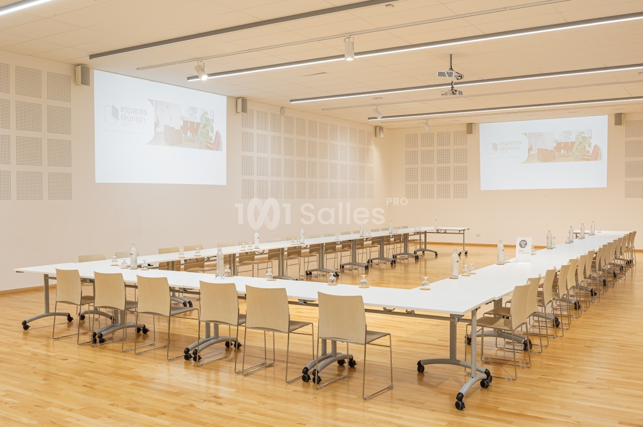Salle de réunion spacieuse avec tables disposées en U, chaises en bois clair et projecteurs au plafond.