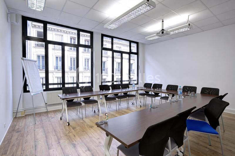 Salle de réunion lumineuse avec tables en bois, chaises noires, paperboard et grandes fenêtres donnant sur des immeubles.