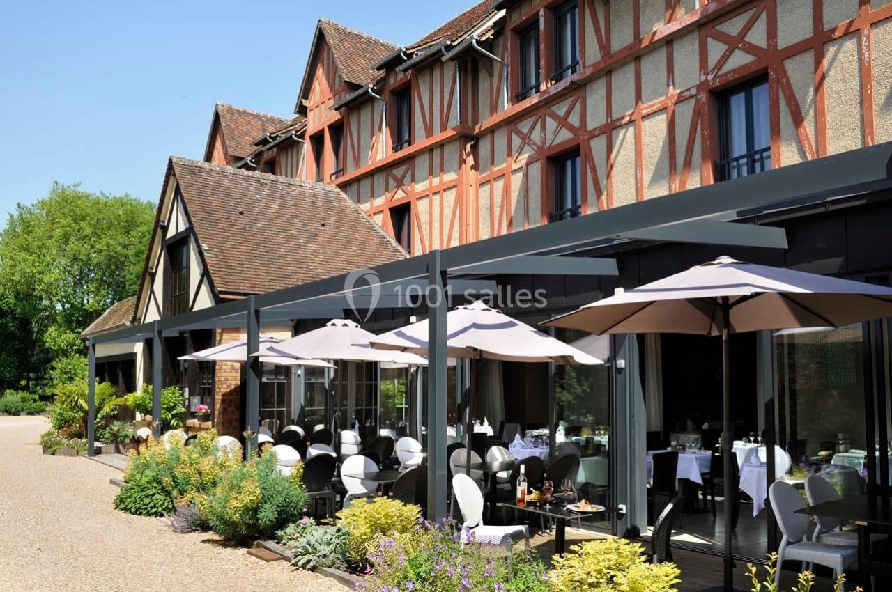 Façade d'un bâtiment à colombages avec une terrasse aménagée, tables dressées et parasols, entourée de verdure.