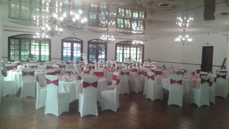 Salle de réception décorée avec des tables rondes, nappes blanches et chaises ornées de nœuds rouges.