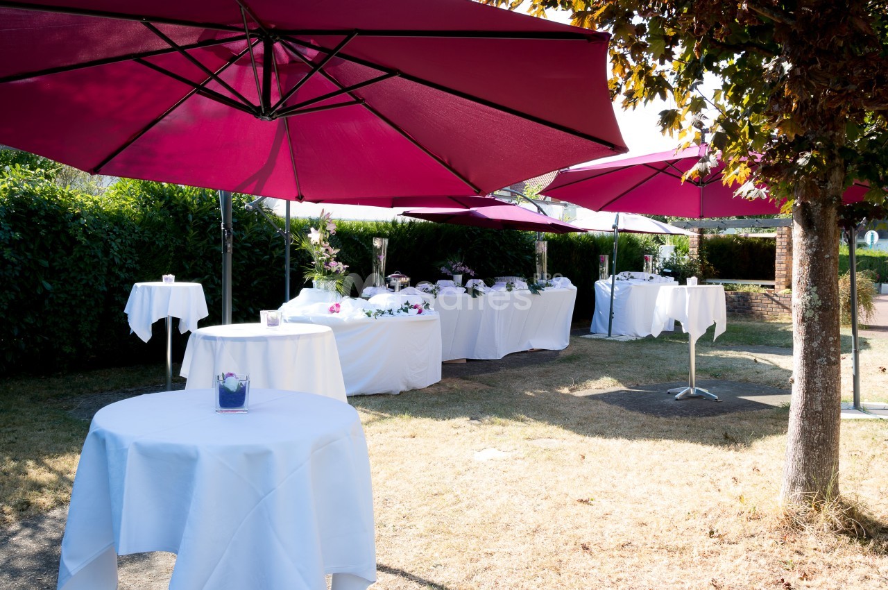 Tables dressées sous des parasols rouges dans un jardin, avec nappes blanches et décorations pour un événement en plein air.