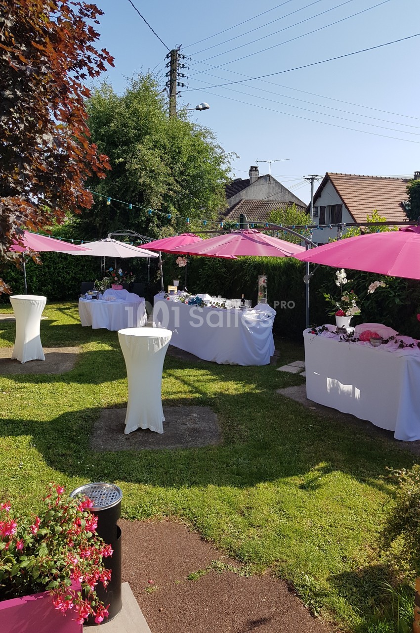 Jardin aménagé pour une réception avec tables décorées, nappes blanches et parasols roses, entouré de verdure.