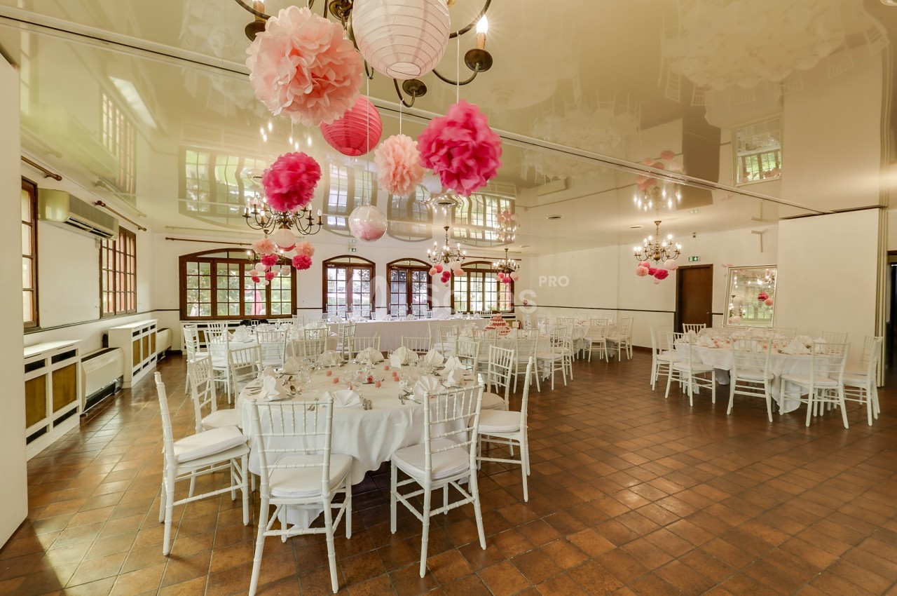 Salle de réception décorée de lanternes et pompons roses, tables rondes dressées avec nappes blanches et chaises assorties.