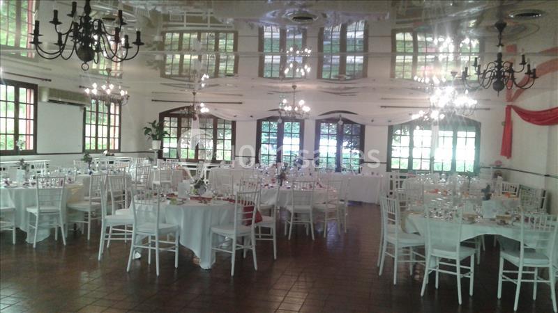 Salle de réception lumineuse avec tables rondes dressées, chaises blanches et lustres suspendus.