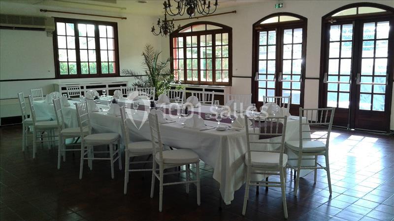Salle lumineuse avec une grande table dressée, nappes blanches, chaises blanches et fenêtres en bois donnant sur l'extérieur.