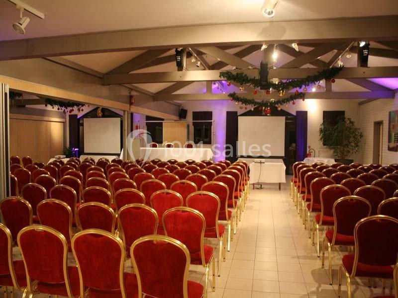 Salle de conférence avec des rangées de chaises rouges alignées face à une table et des écrans de projection.