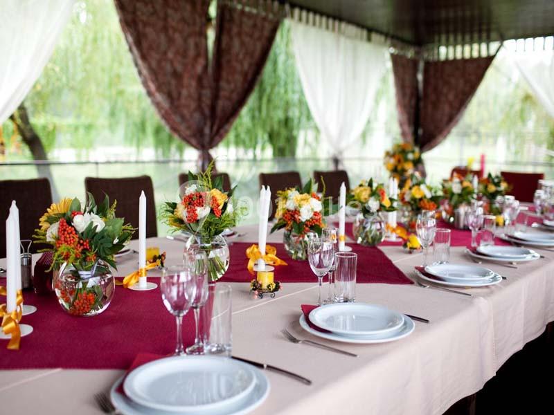 Table dressée avec nappes beige et bordeaux, décorée de bouquets colorés, bougies blanches et vaisselle élégante.