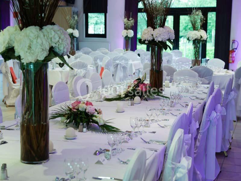 Salle de réception décorée avec des tables dressées, nappes blanches, compositions florales et chaises recouvertes.