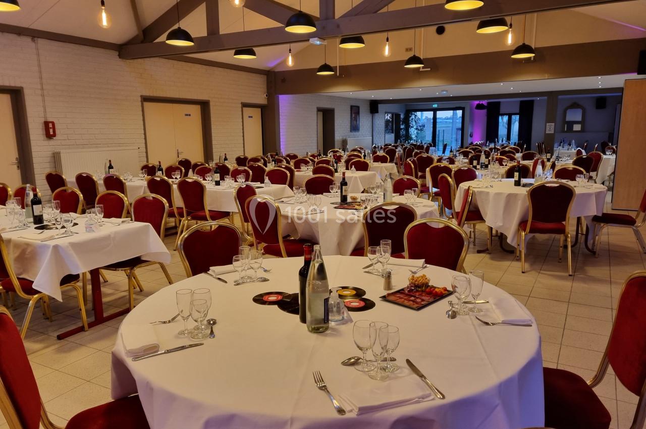 Salle de réception avec tables rondes dressées, nappes blanches, chaises rouges et éclairage suspendu.