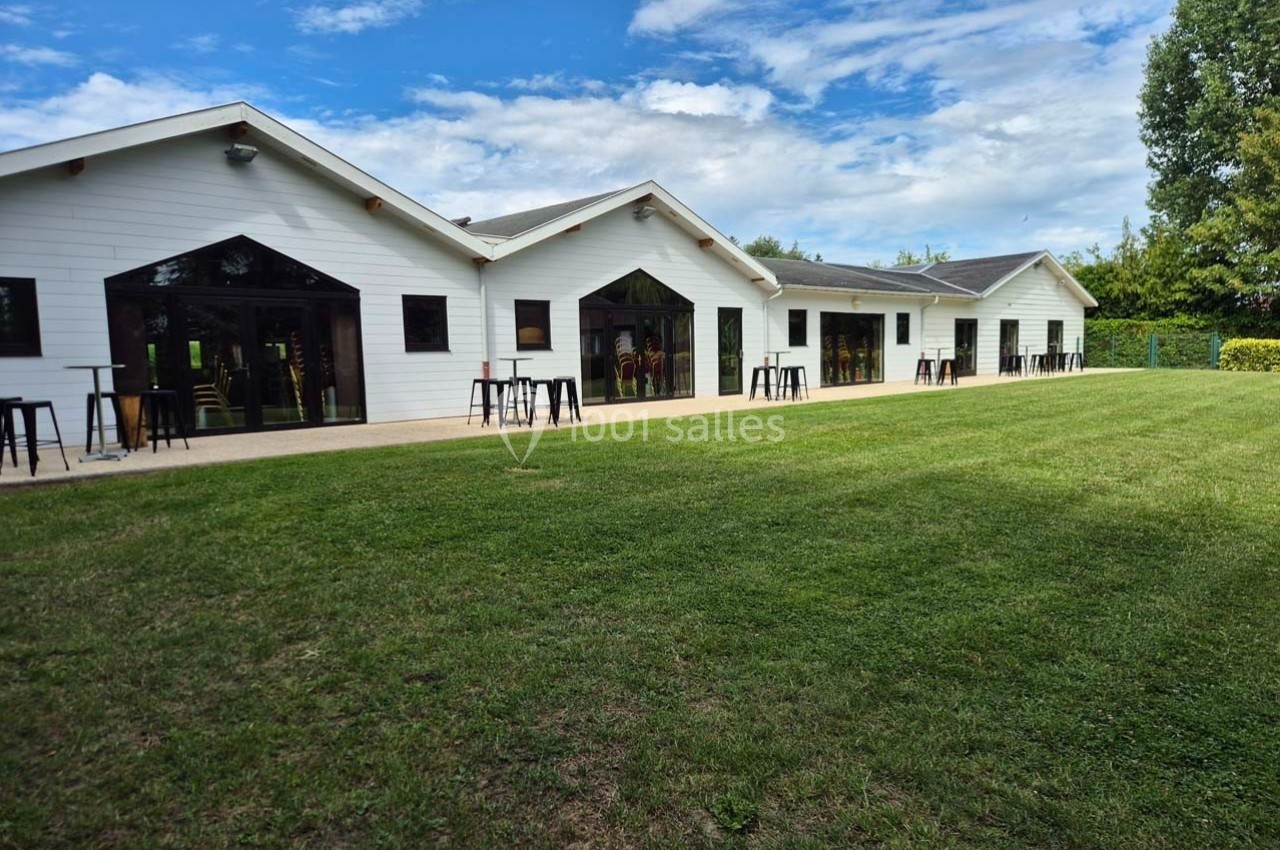 Bâtiment blanc moderne avec grandes baies vitrées, entouré d'une pelouse et d'arbres sous un ciel partiellement nuageux.
