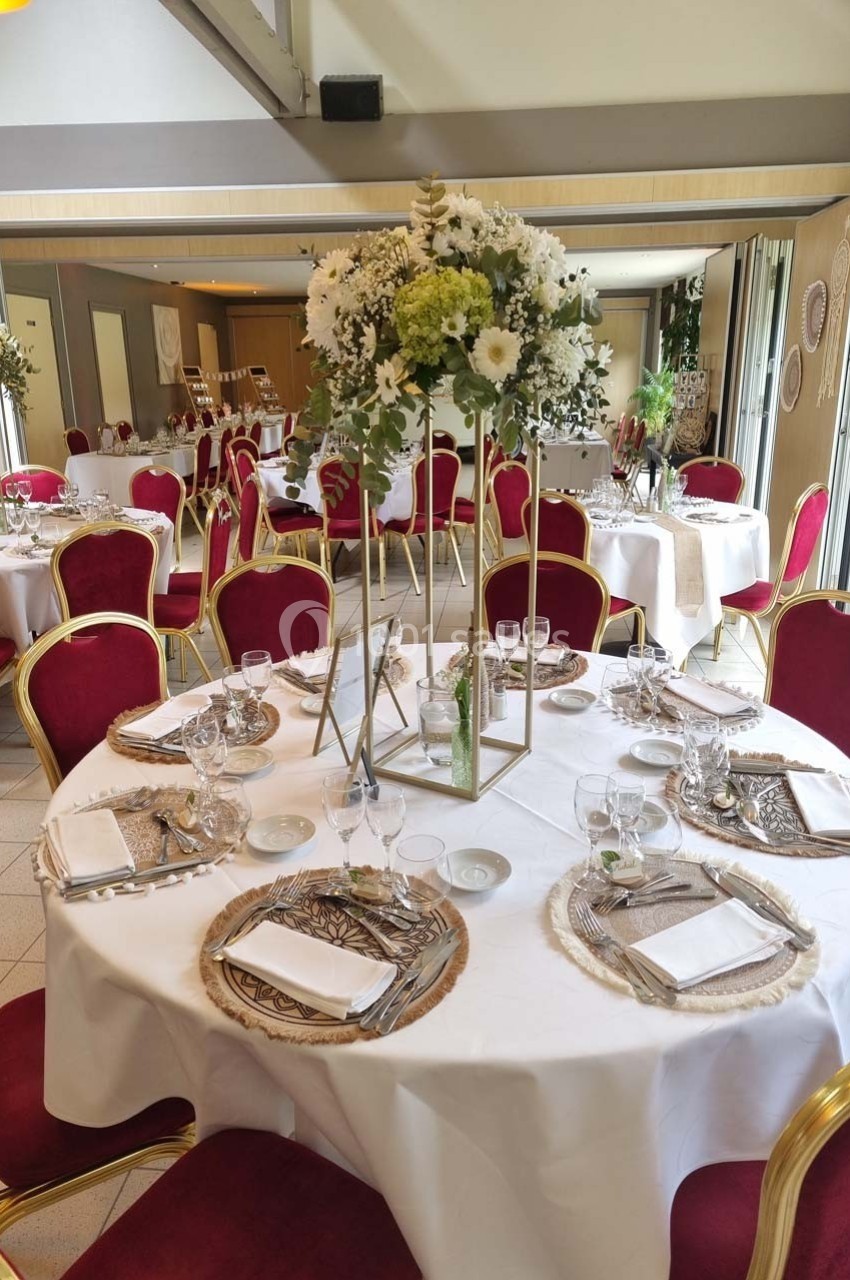 Salle de réception avec tables rondes dressées, nappes blanches, chaises rouges et centre de table floral.