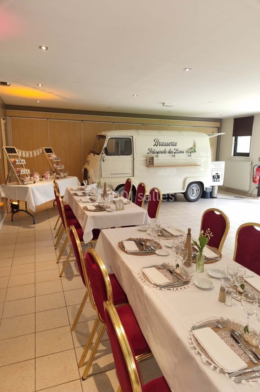 Salle décorée pour un événement avec tables dressées, chaises rouges et un camion rétro blanc en arrière-plan.