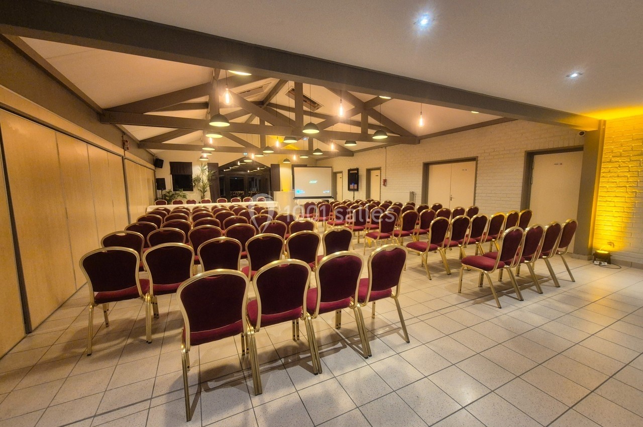 Salle de conférence avec des rangées de chaises rouges alignées face à un écran de projection sous un plafond en poutres…