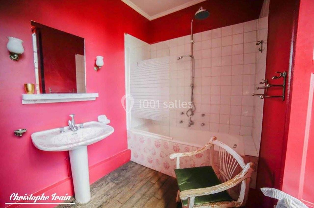 Salle de bain avec murs rouges, baignoire blanche, rideau de douche, lavabo et chaise en bois avec coussin vert.