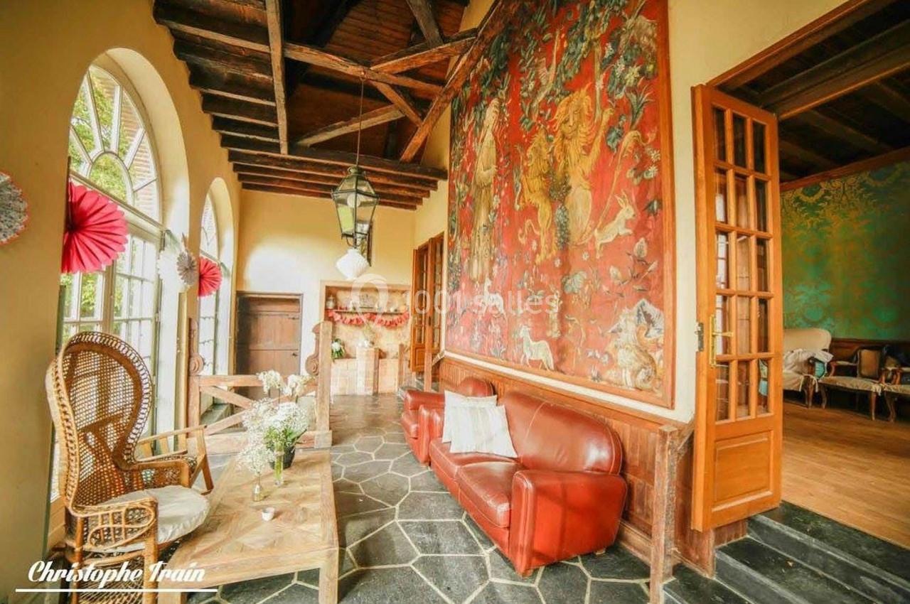 Salon lumineux avec canapé en cuir rouge, fauteuil en osier, grandes fenêtres et fresque murale colorée.