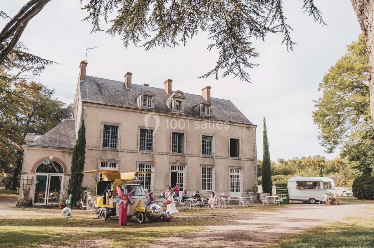 Manoir ancien entouré d'arbres, avec une terrasse aménagée, un food truck jaune et une caravane blanche.