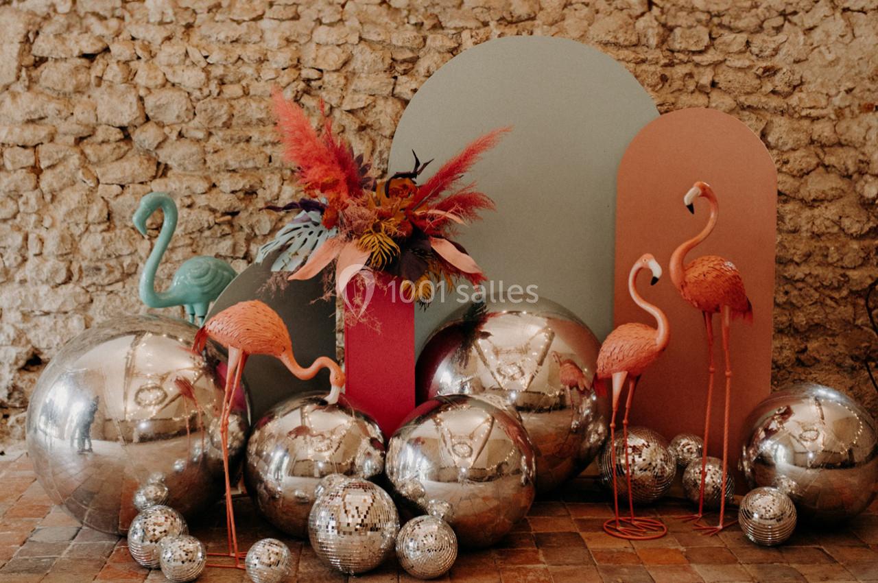Décoration festive avec flamants roses, boules miroir et fleurs colorées devant un mur en pierre.