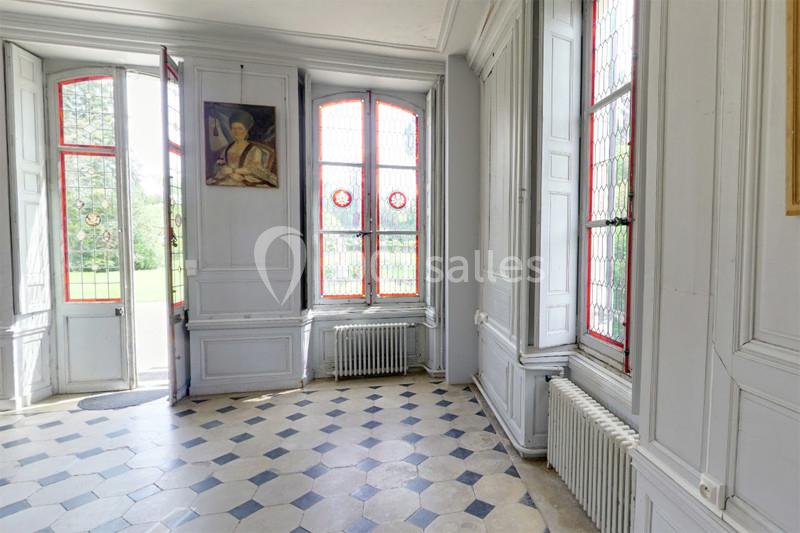 Salle lumineuse avec carrelage en damier, grandes fenêtres à vitraux colorés et un tableau accroché au mur.