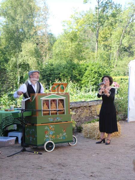 Un homme joue de l'orgue de barbarie tandis qu'une femme chante en plein air, entourés de verdure.